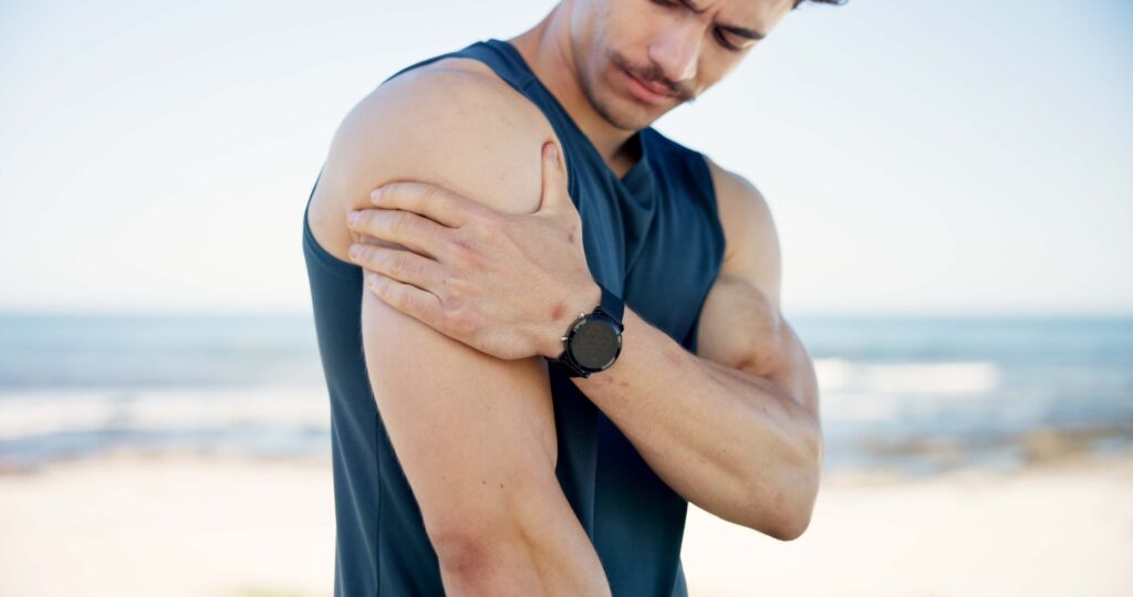Athletic man holding his shoulder while experiencing shoulder pain or discomfort outdoors near the beach.