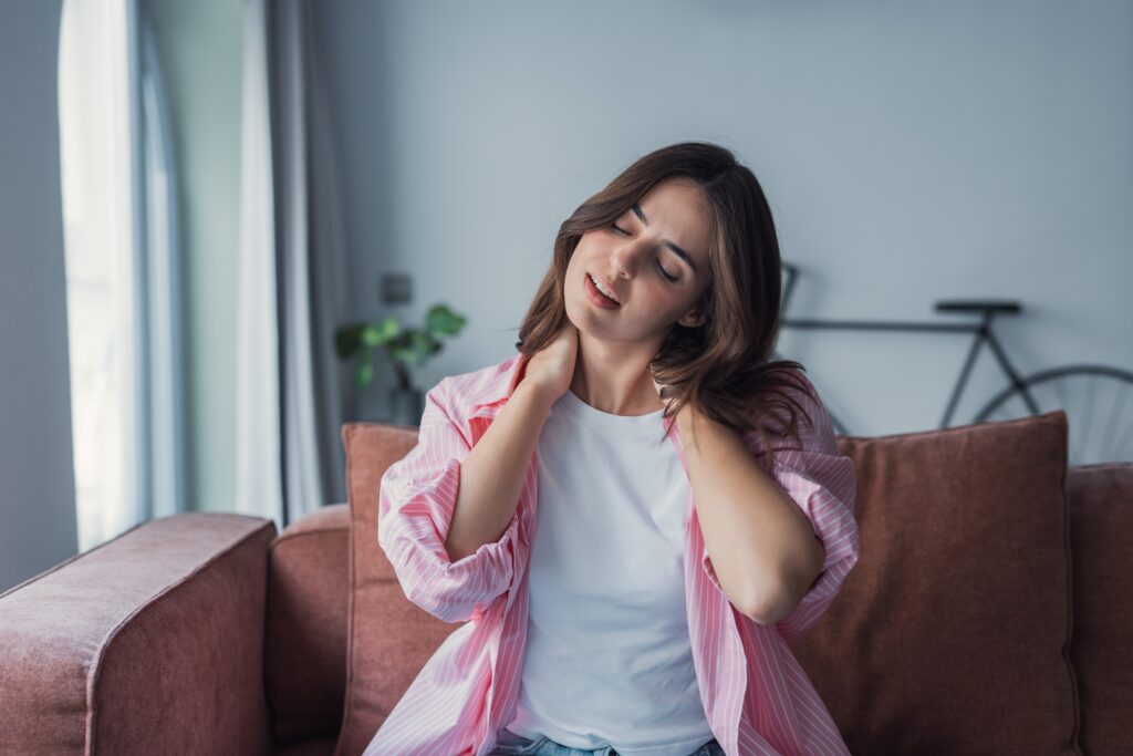 Woman massaging her neck while sitting on a couch, experiencing muscle tension and discomfort from everyday activities.