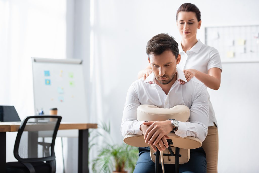 Fully clothed employee receiving a seated shoulder massage in a workplace conference room.